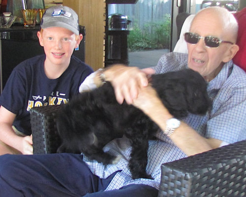 Boy in baseball cap, black puppy being held by older male in sun glasses;2019 Lee's son and father who is holding their puppy.