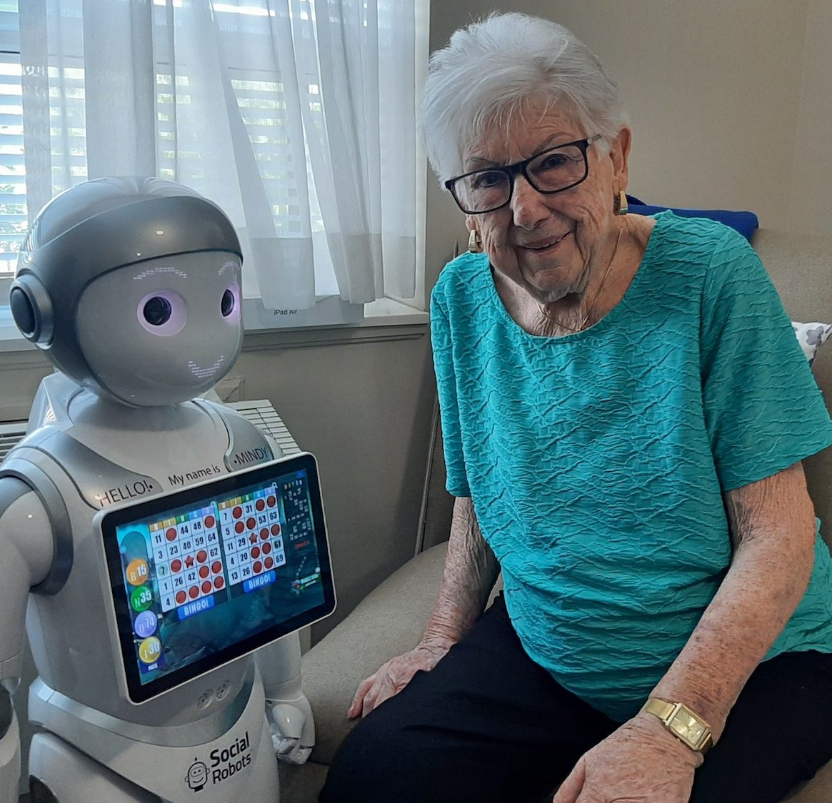 Mindy, robot, with bingo game on chest tablet; standing beside older adult smiling while playing the game.