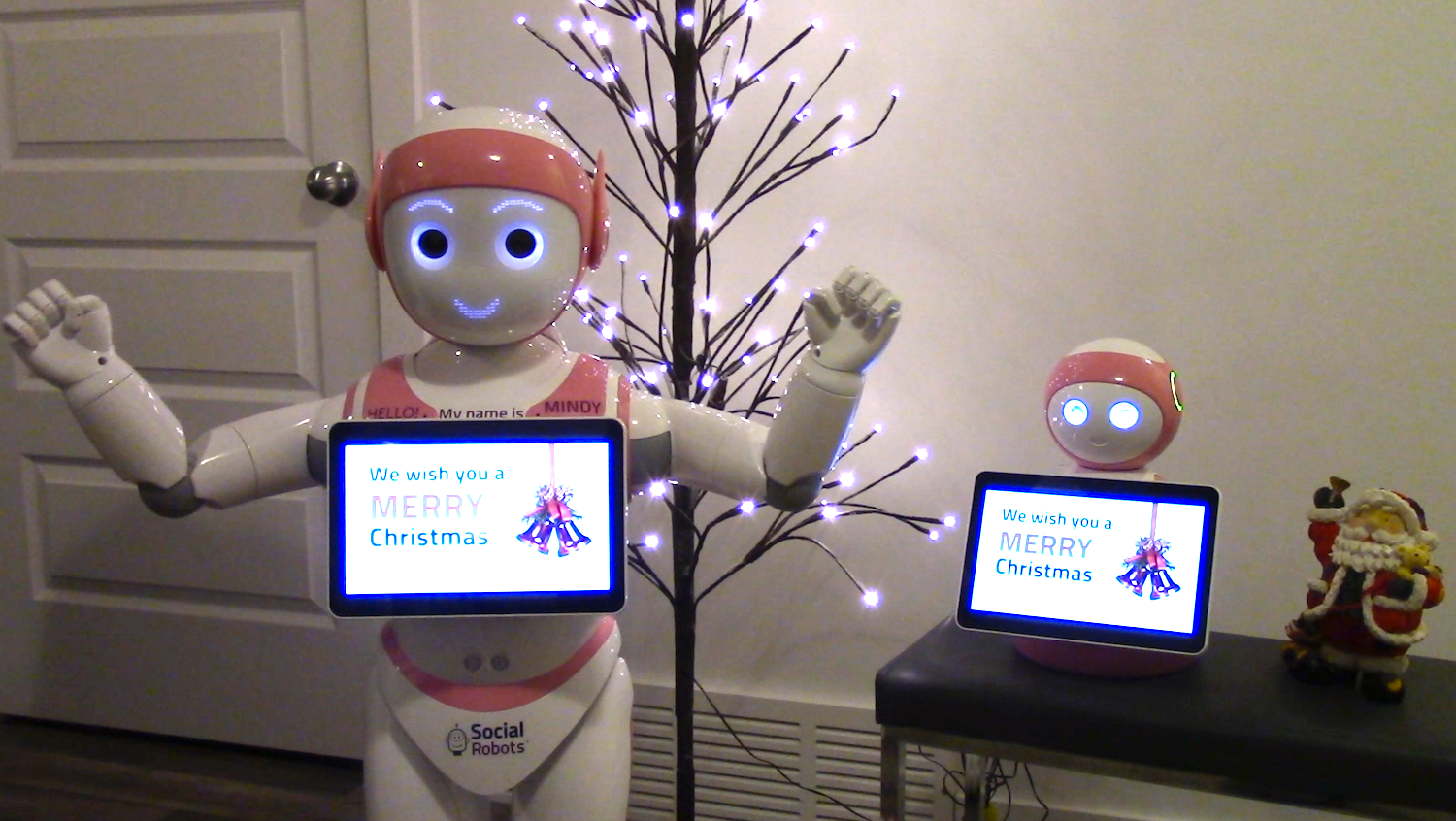 Mindy with arms raised, Merry Christmas song on tablet, mini Mindy to the right side with mini Santa statue